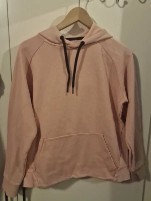Ljusrosa hoodie med svart snöre - En stilren ljusrosa hoodie med huva och svarta dragsnören. Hoodien har en stor magficka framtill och är tillverkad i mjukt material, perfekt för en avslappnad look. Långa ärmar och ribbade muddar vid ärmslut och nederkant.