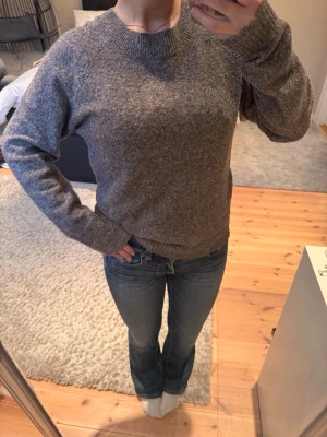 Gråmelerad stickad tröja från Vero Moda  - Mysig gråmelerad stickad tröja med rund halsringning och långa ärmar. Tröjan har en enkel, klassisk design och ribbstickade muddar vid ärmslut och nederkant. Perfekt för kyliga dagar och lätt att matcha med jeans eller kjol.