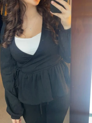 Svart omlottblus med knytband - Snygg svart omlottblus med knytband i midjan och lätt rynkad nederdel. Blusen har långa ärmar och v-ringad halsringning. Perfekt att styla med ett linne under som jag gjort. Helt slutsåld