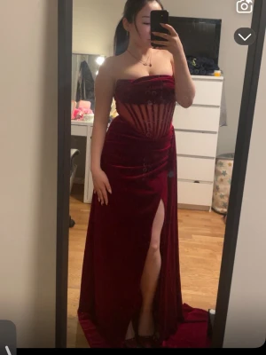 Vinröd långklänning med slits - Säljer en vinröd långklänning med axelbandslös design och markerad midja. Klänningen har en snygg slits framtill och transparenta detaljer i livet som ger en modern look. Perfekt för bal eller festliga tillfällen. Köpte den för ca 300kr. PRIS KAN DISKUTERAS