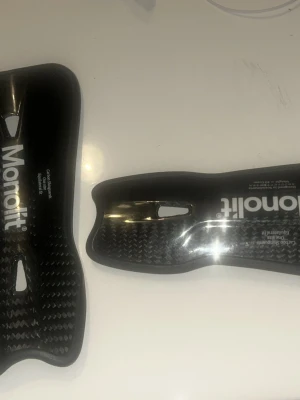 Monolit benskydd i kolfiber - Säljer ett par svarta benskydd från Monolit, tillverkade i lätt och slitstark kolfiber. De har en ergonomisk form med ventilationshål och mjuk kant för extra komfort. Perfekta för dig som vill ha smidiga och tåliga skydd till fotboll eller annan sport. Änvänder inte dom så mycket.