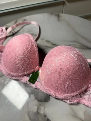 Rosa spets-bh med rosett - Söt rosa bh från Celia Lingerie med vadderade kupor och spetsdetaljer över hela framsidan. Dekorerad med en liten rosett mellan kuporna och justerbara axelband. Perfekt för dig som gillar romantiska detaljer och mjuka material.