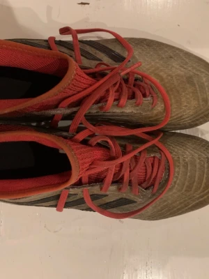 Adidas fotbollsskor predator  - Säljer ett par Adidas fotbollsskor i rött och grått med svarta detaljer. Skorna har snörning och mönstrad ovandel, samt dobbar i rött och svart för bra grepp på planen. Perfekta för dig som vill ha stil och funktion på fotbollsplanen.