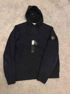 Svart hoodie från Stone Island - Säljer en svart hoodie från Stone Island med klassisk patch på vänster ärm. Hoodien har huva med dragsko och en stor magficka framtill. Tillverkad i mjuk bomull för skön känsla och stilren look. Perfekt för dig som gillar streetwear och exklusiva detaljer.