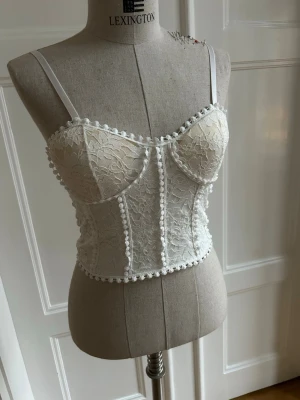 Vit spetskorsett med axelband - Snygg vit korsett i spets med justerbara axelband. Den har en vacker blommig spetsdesign och små dekorativa bollar längs kanterna. Perfekt för att ge en romantisk touch till din outfit.