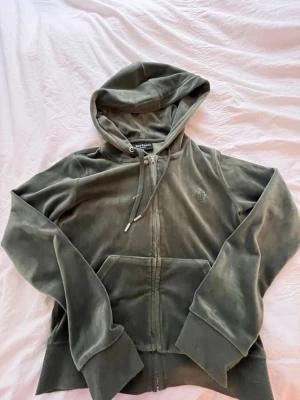 Grön velour hoodie från Juicy Couture - Mysig grön zip-hoodie från Juicy Couture i mjuk velour. Klassisk modell med huva, dragsko och fickor framtill. Broderad JC-logga på bröstet och breda muddar vid ärmslut och nederkant. Perfekt för en chill och trendig look.💚