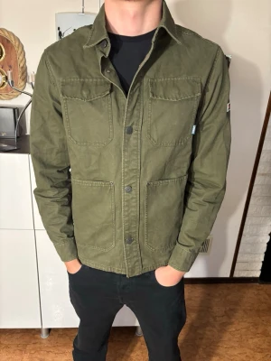 Grön overshirt från Tommy Hilfiger  - Snygg militärgrön overshirt från Tommy Hilfiger med fyra stora fickor framtill och klassisk krage. Tillverkad i bomull och har knappar längs hela framsidan. Perfekt lager-på-lager-plagg för en avslappnad streetstil.
