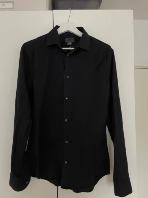 Svart slim fit skjorta från H&M - Klassisk svart skjorta från H&M i slim fit-modell. Skjortan har långa ärmar, knappar framtill och en stilren krage. Perfekt för dig som gillar en enkel och clean look. Tillverkad i mjukt material som känns bekvämt hela dagen.