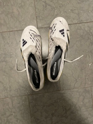 Adidas Predator vita fotbollsskor - Säljer ett par Adidas Predator fotbollsskor i vitt med svarta och gula detaljer. Skorna har klassisk snörning, ränder på sidan och en robust sula med dobbar för bra grepp på planen. Perfekta för dig som vill ha stilrena och funktionella skor till matchen.