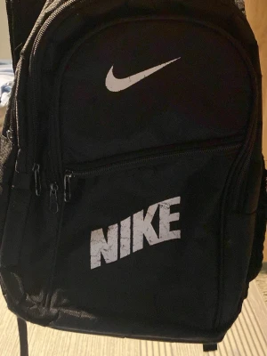 Svart ryggsäck från Nike - Snygg svart ryggsäck från Nike med klassisk logga i vitt. Ryggsäcken har flera fack med dragkedja och vadderad baksida för extra komfort. Perfekt för skola, träning eller utflykter. Tillverkad i slitstark polyester och har justerbara axelband.