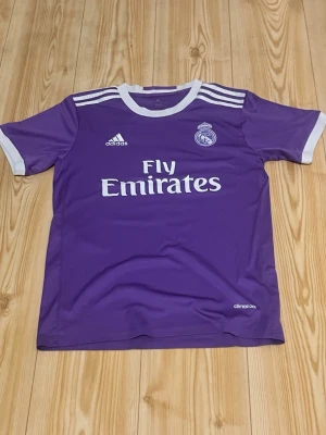 Lila Real Madrid Ronaldo tröja - Snygg lila Real Madrid matchtröja från Adidas med vita detaljer och tryck. Tröjan har Ronaldo 7 på ryggen, klubbmärke och Fly Emirates-logga på bröstet samt klassiska Adidas-ränder på axlarna. Tillverkad i lätt och ventilerande climacool-material.