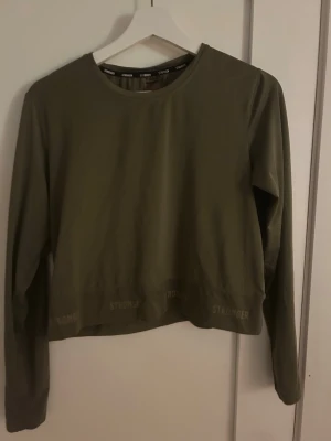 Olivgrön långärmad tröja Stronger - Olivgrön långärmad tröja från Stronger med rund hals och diskret logga längs nederkanten. Tillverkad av 90% återvunnen polyester och 10% elastan, vilket ger en stretchig och bekväm passform. Perfekt för träning eller chill dagar.