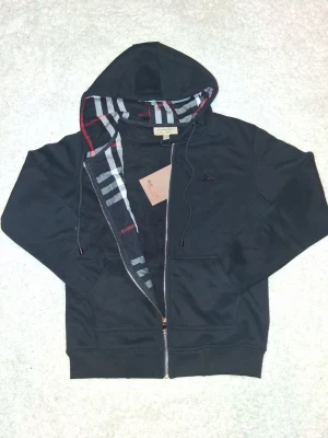 Svart hoodie från Burberry med dragkedja - Svart hoodie från Burberry med dragkedja och klassiskt rutigt foder i huvan. Broderad Burberry-logga på bröstet och justerbar dragsko i huvan. Tillverkad i mjukt bomullsmaterial och har ribbade muddar samt fickor framtill.