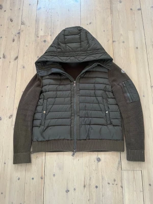 Grön Moncler cardigan  - Snygg grön Moncler cardigan med quiltad/vadderad front och huva i nylon samt stickade ärmar och rygg. Dragkedja framtill, två framfickor med knapp och en ärmficka med dragkedja. Klassisk Moncler-logga på ena ärmen som ger en exklusiv look. Mycket bra skick förutom ett litet hål som syns på bilderna, vilket är inräknat i priset. Passar dig som vanligtvis bär storlek M. Jackan är perfekt nu för lite kyligare väder. Vid frågor eller önskemål om fler bilder är det bara att höra av sig. Mvh Olof.