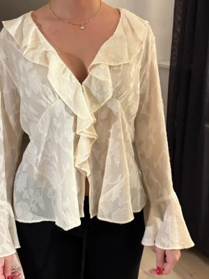 Vit volangblus med blommönster - Säljer en romantisk vit blus med volangkant och blommigt mönster. Blusen har djup v-ringning, transparent tyg och långa, utsvängda ärmar. Perfekt för dig som gillar en feminin och trendig stil. Materialet känns lätt och luftigt.