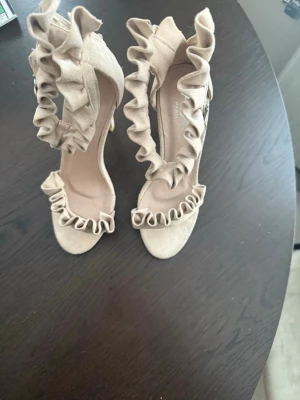 Beige pumps med volang från AX Paris - Snygga beige pumps från AX Paris med hög klack och volangdetaljer både framtill och runt ankeln. Skorna har öppen tå och spänne vid sidan. Tillverkade i mockaimitation för en mjuk och lyxig känsla.