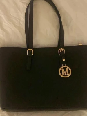 Michael kors liknande - Stilren svart handväska liknade Michael Kors med guldfärgade detaljer och ett stort MK-hänge framtill. Väskan har två handtag och medföljande axelrem med guldfärgade spännen. Perfekt för dig som gillar klassisk och elegant design.