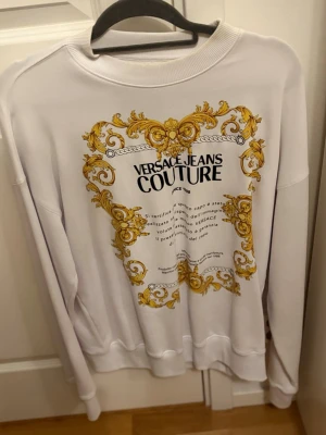 Vit sweatshirt Versace Jeans Couture - Vit sweatshirt från Versace Jeans Couture med stort tryck i svart och guld på ryggen. Tröjan har rund halsringning och långa ärmar. Detaljerad barockinspirerad design i gult och guld ger en lyxig känsla. Perfekt för dig som gillar statement-plagg. (Oversized fit)