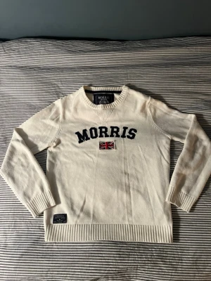 Vit stickad tröja från Morris - Snygg vit stickad tröja från Morris med svart broderad logga och en liten brittisk flagga på bröstet. Tröjan har rund halsringning, ribbade muddar och nederkant. Perfekt för dig som gillar klassisk preppy stil.