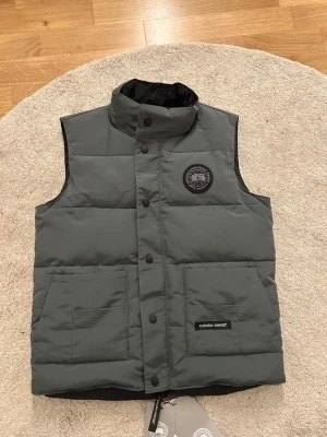 Mörkgrå dunväst från Canada Goose - Snygg mörkgrå dunväst från Canada Goose med hög krage, två stora framfickor och svart logotypmärke på bröstet. Västen har tryckknappar framtill och är perfekt för lager-på-lager under kyliga dagar. Klassisk och stilren design i slitstarkt material.