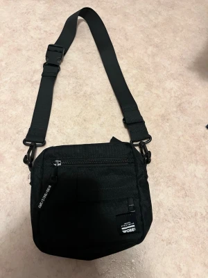 Svart axelväska från Woxit - Snygg svart axelväska från Woxit med justerbar rem och praktiska fack framtill. Väskan har dragkedja och är tillverkad i slitstark polyester, perfekt för att bära det viktigaste. Enkel och stilren design som passar till det mesta.