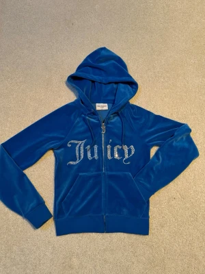 Blå velour hoodie Juicy Couture, Nyskick - Blå zip-up hoodie från Juicy Couture i mjuk velour med huva och fickor framtill. Framsidan har stora glittriga strasstenar med 'Juicy' och dragkedjan har ett J-format hänge. Perfekt för dig som gillar en lyxig och avslappnad stil. Mycket fint skick, knappt använd. Nypris 1199kr.