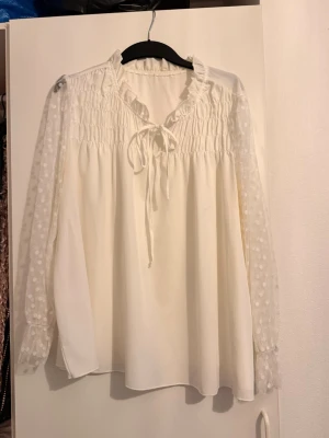 Vit blus med prickiga ärmar och volang - Söt vit blus med transparenta, prickiga långärmar och volangkantad krage. Blusen har smock upptill och knytband vid halsen för en romantisk look. Perfekt till jeans eller kjol för en trendig outfit.