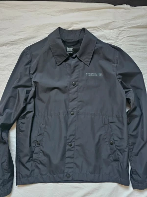 Svart vindjacka från Helmut Lang - Stilren svart vindjacka från Helmut Lang med tryckknappar framtill och två snedställda fickor. Jackan har klassisk krage och diskret logga på bröstet. Ingen prislapp kvar, men aldrig använd, alltså helt ny.