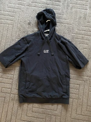 Svart EA7 hoodie med kort ärm - Svart hoodie från EA7 Emporio Armani med korta ärmar och huva med dragsko. Snygg logga på bröstet och två fickor framtill. Tillverkad i mjukt bomullsmaterial, perfekt för en avslappnad och trendig stil.