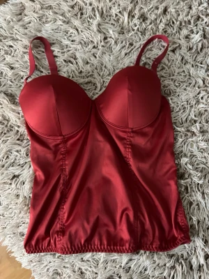 Röd satin korsett med bygel - Snygg röd korsett i glansigt satinmaterial med vadderade kupor och bygel för extra stöd. Justerbara axelband och hakknäppning bak. Perfekt för att skapa en feminin siluett och ge outfiten en extra touch. Ca S i storlek 