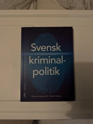 Svensk kriminalpolitik kursbok - Kursbok i svensk kriminalpolitik av Robert Andersson och Roddy Nilsson. Boken har ett blått omslag med ett stort fingeravtryck och tydlig vit text. Perfekt för dig som pluggar juridik, kriminologi, samhällsvetenskap eller politik.