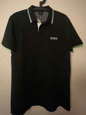 Svart pikétröja från BOSS - Snygg svart pikétröja från BOSS med vit logga på bröstet och ryggen. Kragen har gröna och vita detaljer och ärmarna har matchande gröna kanter. Klassisk passform och korta ärmar, perfekt till en clean och sportig look.