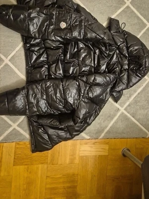 Svart dunjacka från Moncler - Snygg svart dunjacka från Moncler med glansig finish och quiltad design. Jackan har huva, dragkedja och en ficka med Moncler-logga på ärmen. Perfekt för kalla dagar och ger en riktigt clean look.