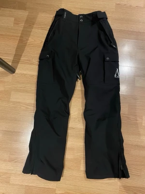 Svarta täckbyxor från Slade stl L - Svarta täckbyxor från Slade med raka ben, praktiska cargofickor på sidorna och dragkedjor vid bensluten. Byxorna har justerbar linning.