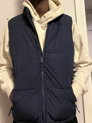 Mörkblå dunväst med dragkedja - Stilren mörkblå dunväst med hög krage och hel dragkedja framtill. Västen har två sidofickor och är lätt vadderad för extra värme. Passar perfekt över en hoodie för en avslappnad look. Materialet är glansigt på insidan och matt på utsidan.