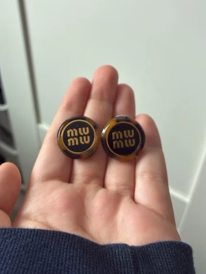 Oäkta Miu Miu runda örhängen i brun plast - Säljer ett par runda örhängen från Miu Miu i brunmelerad plast med logga i guld på framsidan. De har en cool retrovibe och fästs med klassisk stiftbaksida. Perfekta för dig som gillar statement-accessoarer och vill sticka ut lite extra.