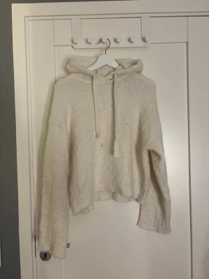  stickad hoodie med luva - Mysig stickad hoodie i offwhite med luva och dragsko. Tröjan har avslappnad passform, långa ärmar och ribbade kanter. Perfekt för lager-på-lager och chill dagar. Vill påpeka att den har ett litet hål vid halsen och liten fläck där nere vid magen ungefär!!