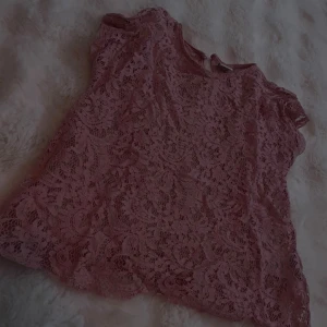 Rosa spetsblus med kort ärm - Söt rosa blus i spets med korta ärmar och rund halsringning. Blusen har ett genombrutet spetsmönster och en liten knappdetalj i nacken. Perfekt för dig som gillar romantisk och feminin stil. Står ingen storlek men hyfsat liten 