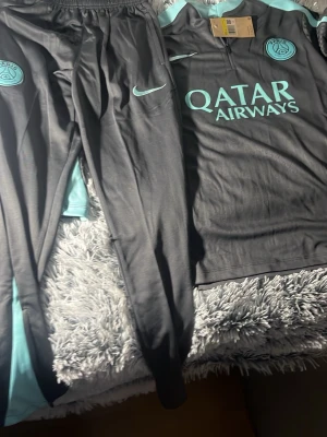 PSG träningsjacka Nike svart/turkos - Snygg långärmad tränings set från PSG i grå med turkosa detaljer och loggor. Jackan har halv dragkedja framtill, PSG-märke på bröstet och Qatar Airways-tryck. Perfekt för fotbollsträning eller chill. Tillverkad i lätt och funktionellt material.