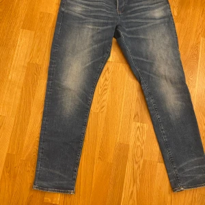 Blå jeans från G-Star RAW - Säljer ett par klassiska blå jeans från G-Star RAW med snyggt slitna detaljer och raka ben. Jeansen har fem fickor och en brun patch med logga bak i midjan. Perfekta för en avslappnad och trendig look.