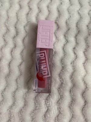 Maybelline Lifter Plump Läppglans - Säljer ett Maybelline Lifter Plump läppglans med rosa lock och transparent hylsa som visar ett glansigt, rosa läppglans. Förpackningen har en modern design med tydlig logga och text. Perfekt för dig som vill ha fylligare och glansiga läppar.