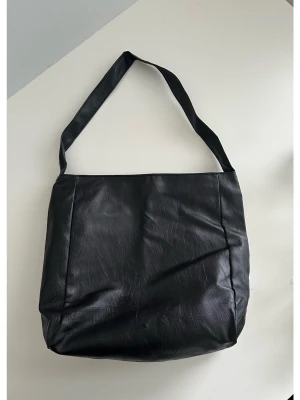 Svart axelväska i skinnimitation - Stilren svart axelväska i skinnimitation med enkel design och bred axelrem. Väskan har en mjuk, lite slouchy form och passar perfekt för dig som vill ha plats för allt du behöver under dagen. Inga synliga loggor eller detaljer, bara clean och klassisk look.