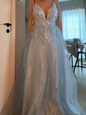 Ljusblå långklänning med spetsdetaljer - Magisk ljusblå långklänning med tunna axelband, djup v-ringning och glittrande tyll. Klänningen har vackra spetsapplikationer och blommiga detaljer upptill. Perfekt för bal eller student. Flödande kjol i flera lager ger en sagolik känsla.