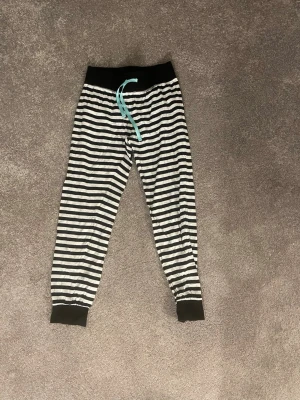 Randiga pyjamasbyxor med snöre, barnstorlek, 134-140/8-10y - Mysiga pyjamasbyxor med svartvita horisontella ränder och svart midjeresår. Byxorna har ett turkost dragsnöre i midjan och muddar vid bensluten. Perfekta för chillkvällar hemma.