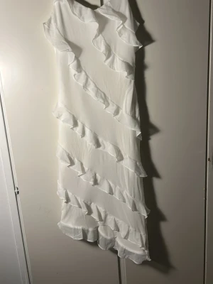 Vit klänning med volanger – populär studentklänning - Vit klänning med volanger och asymmetrisk nederkant. Ärmlös och trendig – använd endast en gång. Perfekt som studentklänning eller till fest! 