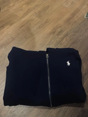 Marinblå zip hoodie från Polo Ralph Lauren - Snygg marinblå hoodie med dragkedja från Polo Ralph Lauren. Klassisk design med vit broderad logga på bröstet och mjukt material. Perfekt för en clean och stilren look. Passar till det mesta och är enkel att matcha passar även M. Mer frågor kom privat😃