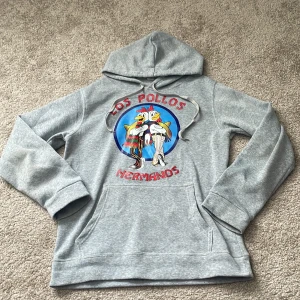Grå Los Pollos Hermanos hoodie - Säljer en grå hoodie med stor Los Pollos Hermanos-logga på bröstet. Hoodien har huva med dragsko och en klassisk magficka. Tillverkad i mjukt sweatshirtmaterial, som tunn fleece, perfekt för en avslappnad streetwear-look.