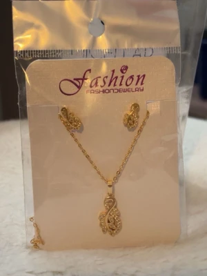 Guldfärgat smyckeset med halsband - Snyggt smyckeset i guldfärg med halsband och matchande örhängen. Både halsbandet och örhängena har ett glittrande, snirkligt mönster med små stenar. Perfekt för att lyfta din outfit med lite extra bling.