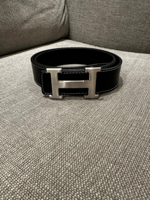 Svart H bälte med H-spänne - Snyggt svart bälte från H med ikoniskt H-format metallspänne. Bältet är gjort i slätt läder med vita kontrastsömmar längs kanterna. Perfekt accessoar för att lyfta vilken outfit som helst med en lyxig touch.