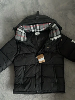 Svart burberry jacka  - Snygg svart dunjacka  med rutigt foder i huvan och dragkedja framtill. Jackan har två framfickor med dragkedja och tryckknappar, samt en klassisk pufferdesign som håller dig varm under kalla dagar. Storleken är S men sitter som xs 